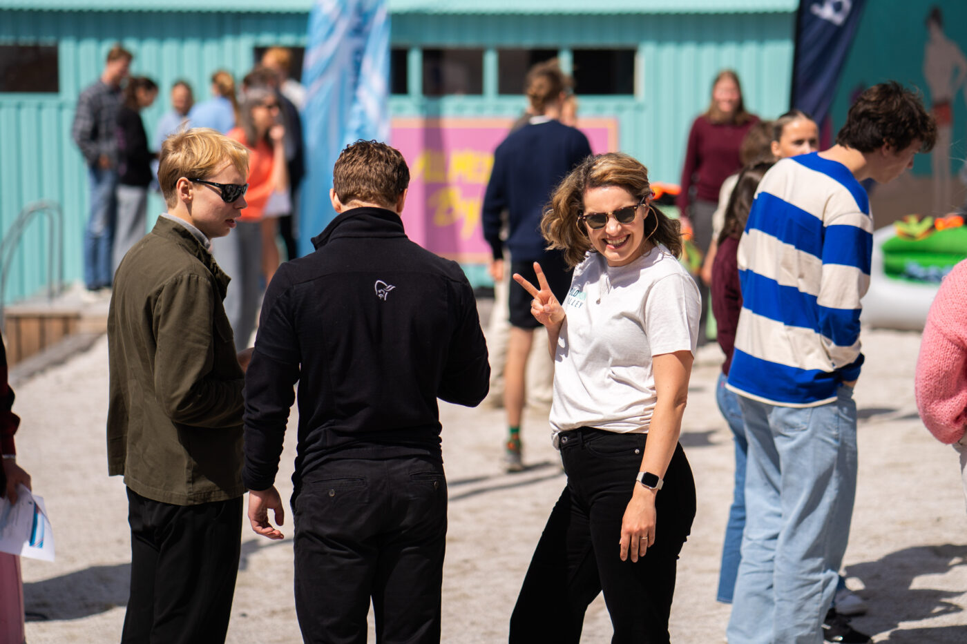 Kvinne med solbreiller holder opp peace-tegn på en bystrand med andre mennesker.